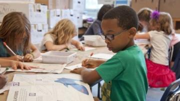 A little boy looks at a book while his classmates are writing.