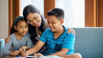 A mom helps her son with homework while the daughter watches.